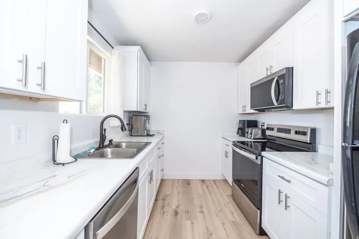 Galley kitchen featuring white cabinetry, light wood-finish flooring, stainless steel appliances, a double basin sink with a matte black faucet, and white countertops with subtle veining