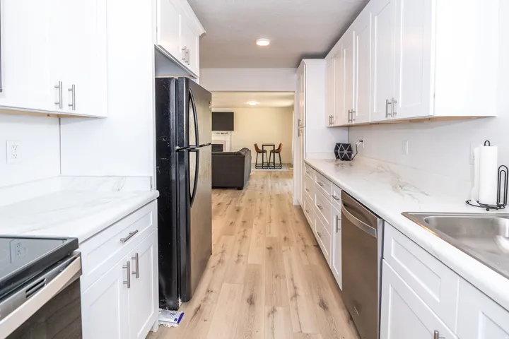 Galley kitchen featuring light wood-finish flooring, white shaker cabinetry, white countertops with subtle veining, and a stainless steel dishwasher