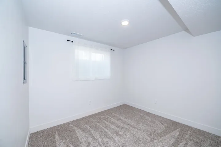 Carpeted interior room featuring white walls, a single window with sheer white curtains, a white ceiling with a recessed light, and a visible electrical panel