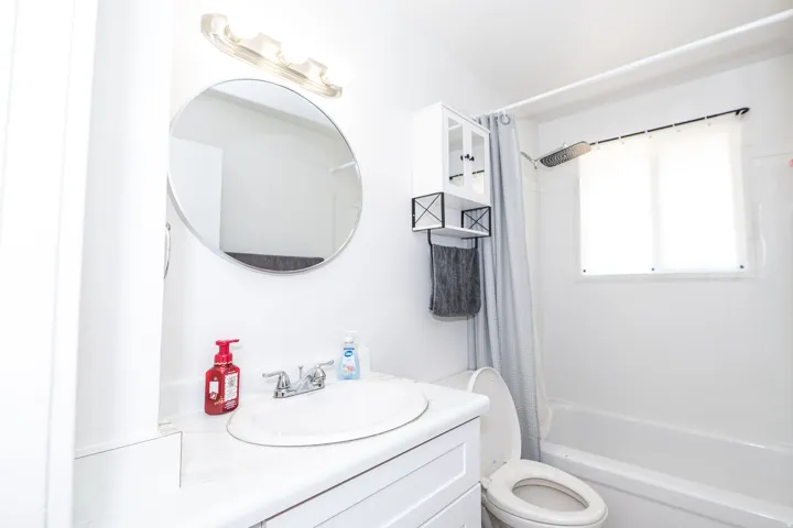 Bathroom featuring a single vanity with a white countertop and integrated sink, a round wall-mounted mirror, and a built-in bathtub with a shower