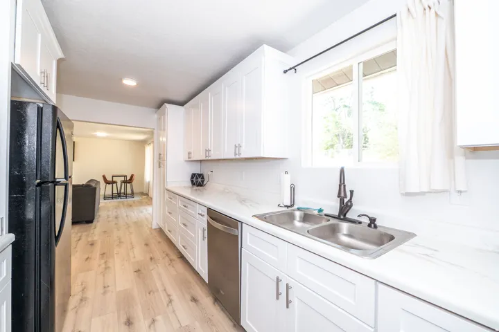 Bright kitchen featuring white shaker cabinetry, stainless steel double-basin sink, bronze-finish faucet, white marble-look countertops, and wood-finish flooring