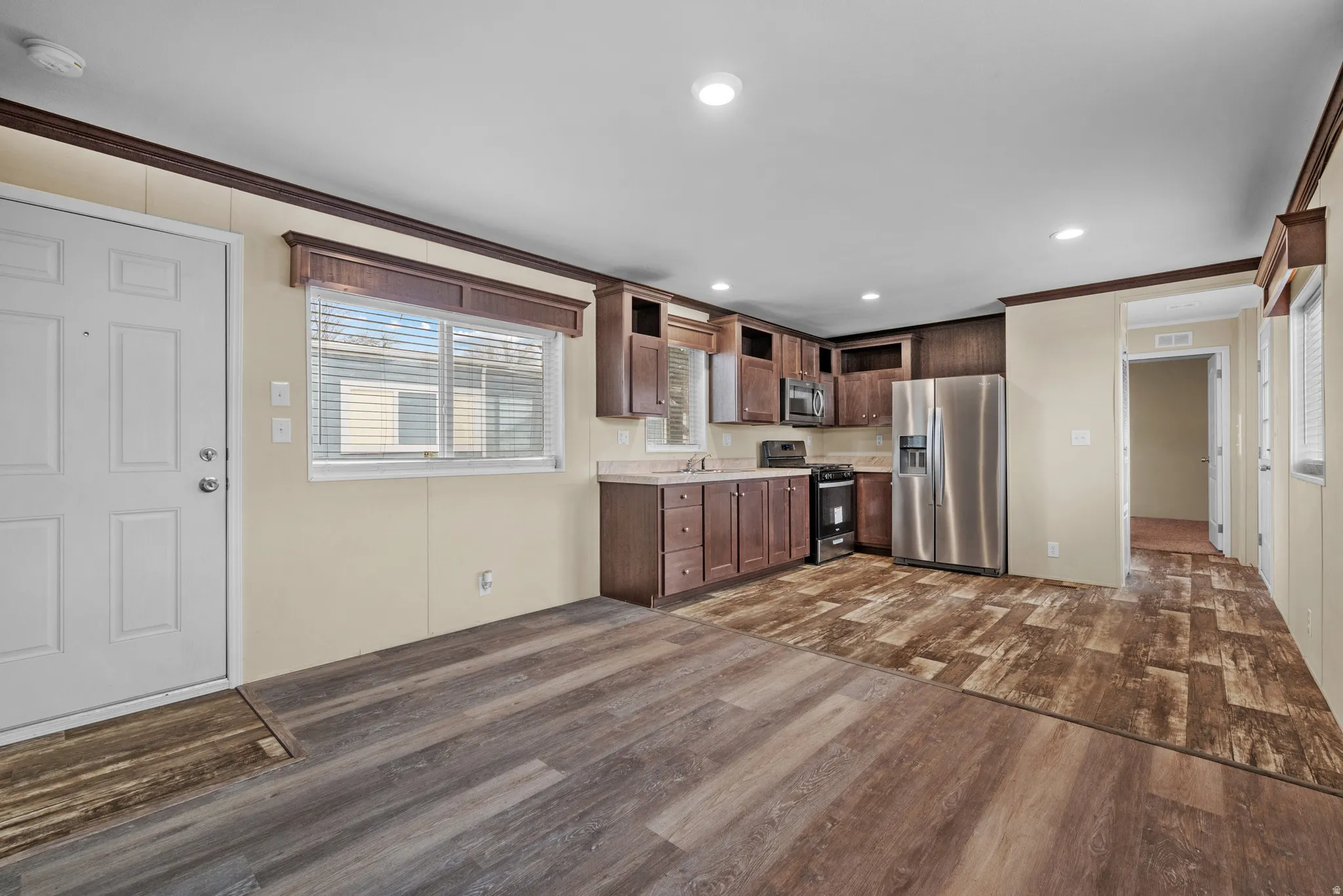 Open-concept kitchen and living area featuring wood-finish flooring, recessed lighting, dark wood cabinetry, stainless steel appliances, and a window with blinds