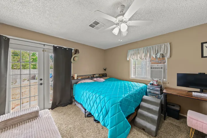 Bedroom with direct outdoor access via double French doors, featuring a ceiling fan with integrated lighting, a window with an installed AC unit, and textured carpeting