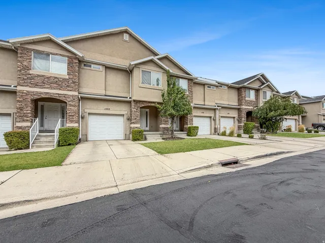 Multi-story townhome exterior featuring an attached garage, stone veneer accents, stucco siding, and a covered entry with white railings