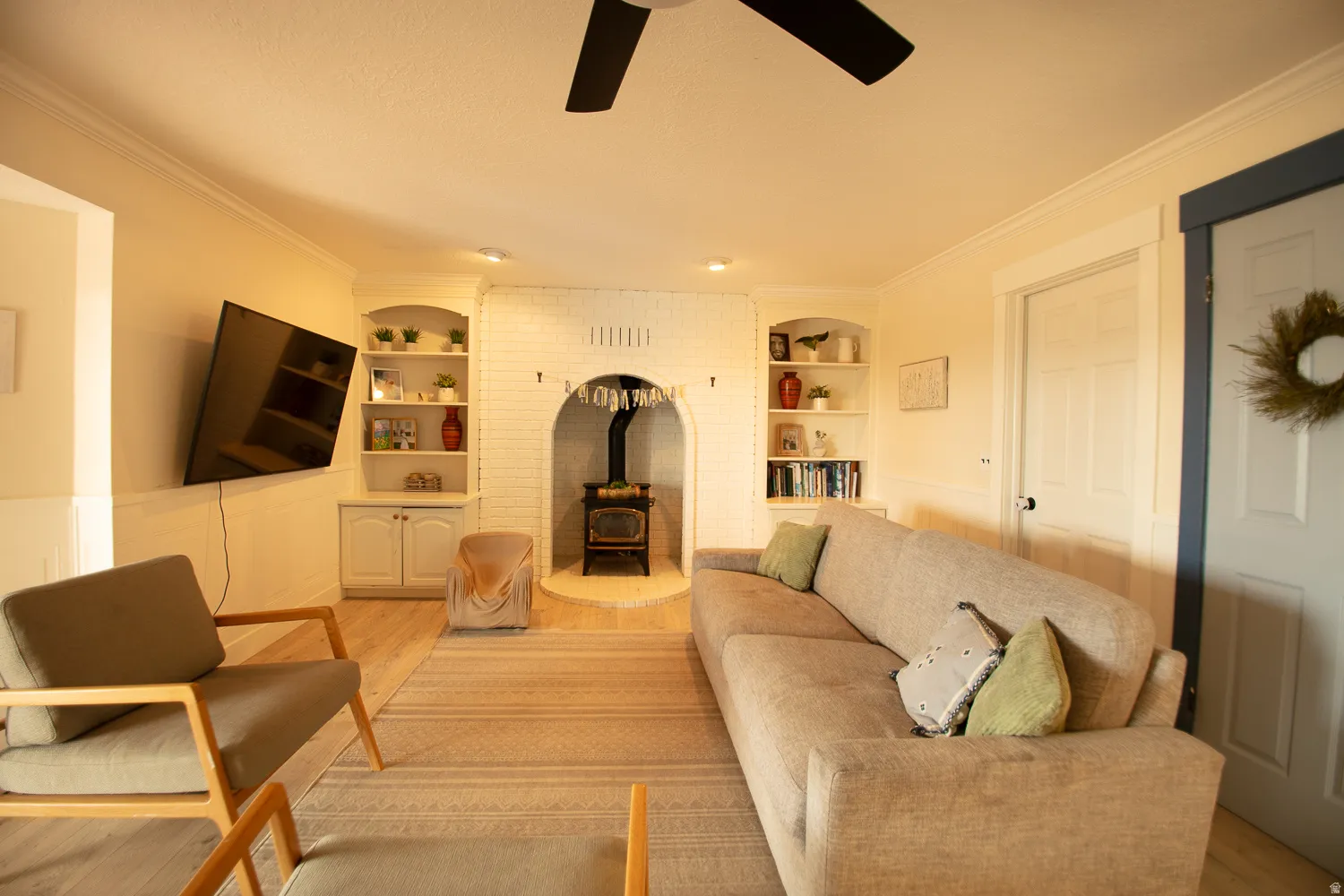 Living area featuring a white-painted brick fireplace with a wood-burning stove, built-in shelving, wood-finish flooring, and crown molding