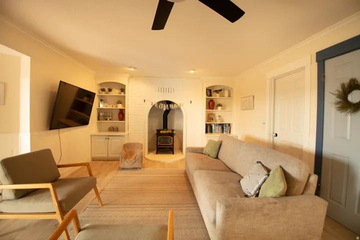 Living area featuring a white-painted brick fireplace with a wood-burning stove, built-in shelving, wood-finish flooring, and crown molding