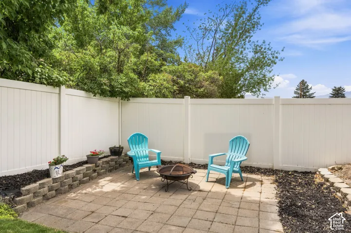 Paved patio area featuring a fire pit, bordered by a white privacy fence and mature trees