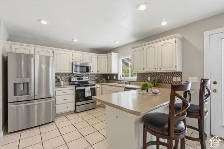 Kitchen featuring stainless steel appliances, light-toned cabinetry, herringbone-patterned backsplash, recessed lighting, and a breakfast bar