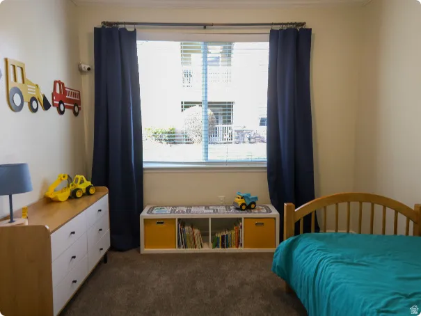 Carpeted room featuring a window with blinds and dark curtains, light beige walls, and a built-in storage unit with open shelving