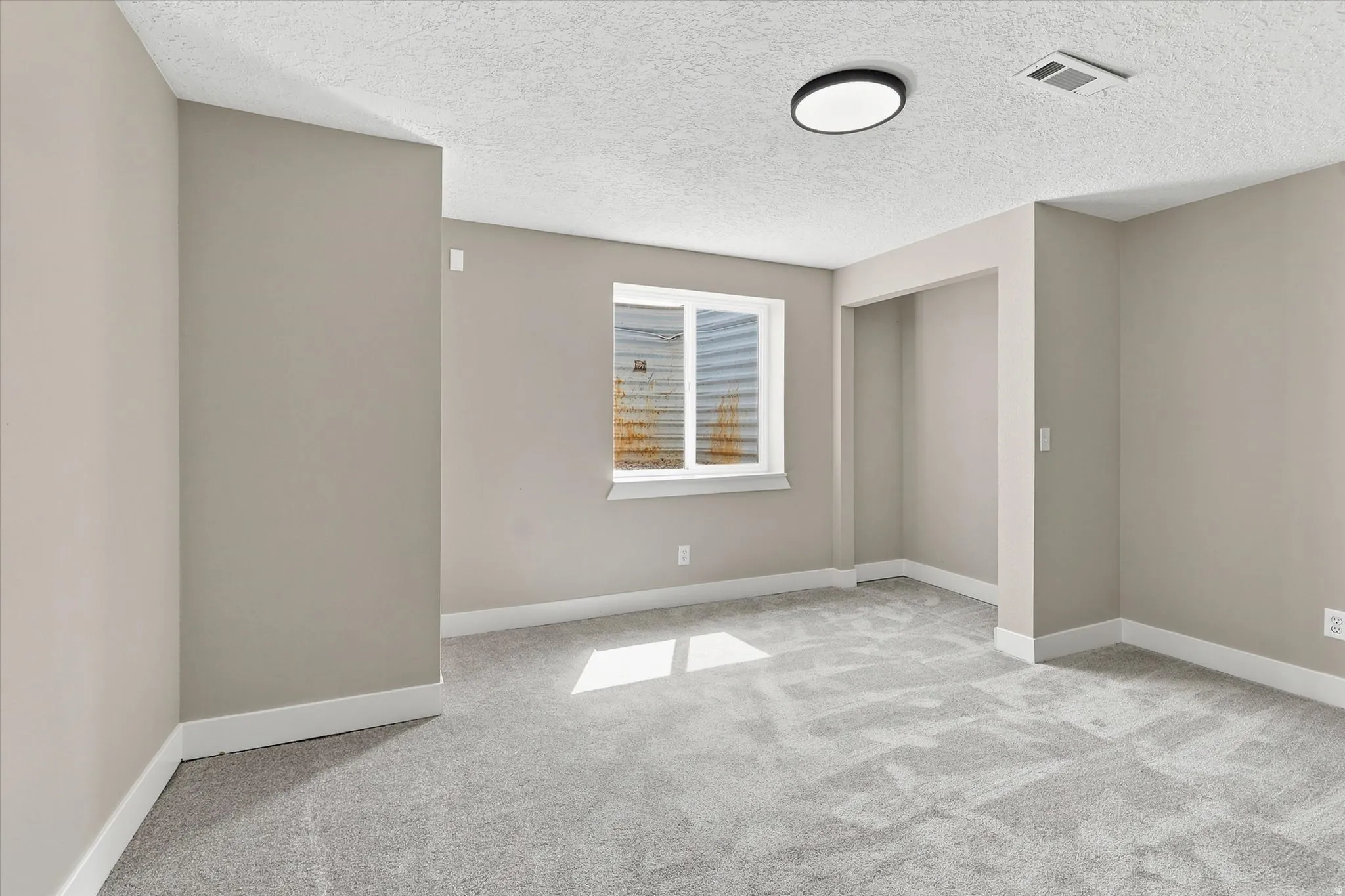 Spacious room featuring neutral paint, gray carpeting, a single window, white baseboards, and an overhead flush-mount light fixture