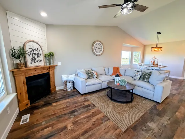 Vaulted ceiling with a ceiling fan, wood-finish flooring, corner fireplace with a wood mantel, and bay windows