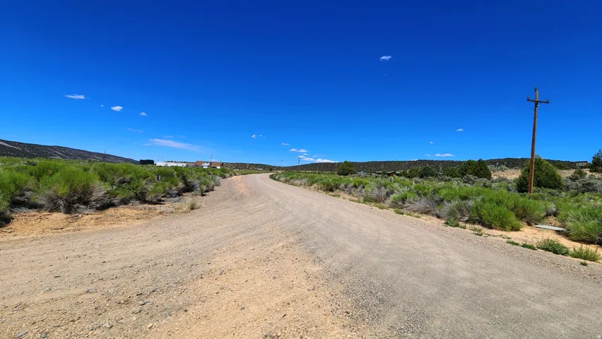 Expansive unpaved road with surrounding natural shrubbery, leading towards distant structures under a clear sky