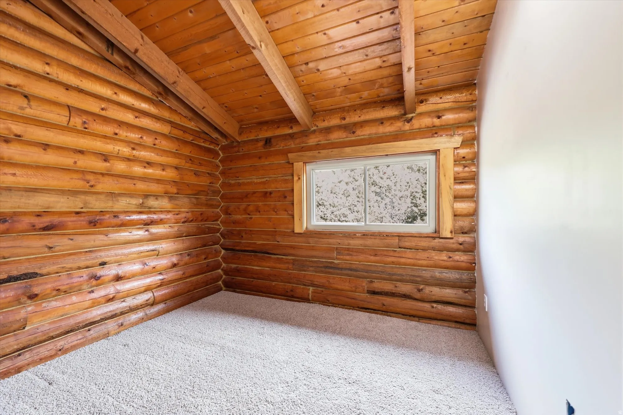 Interior room featuring log walls, an exposed beam ceiling, a window with natural wood trim, a light-colored carpet, and a painted wall