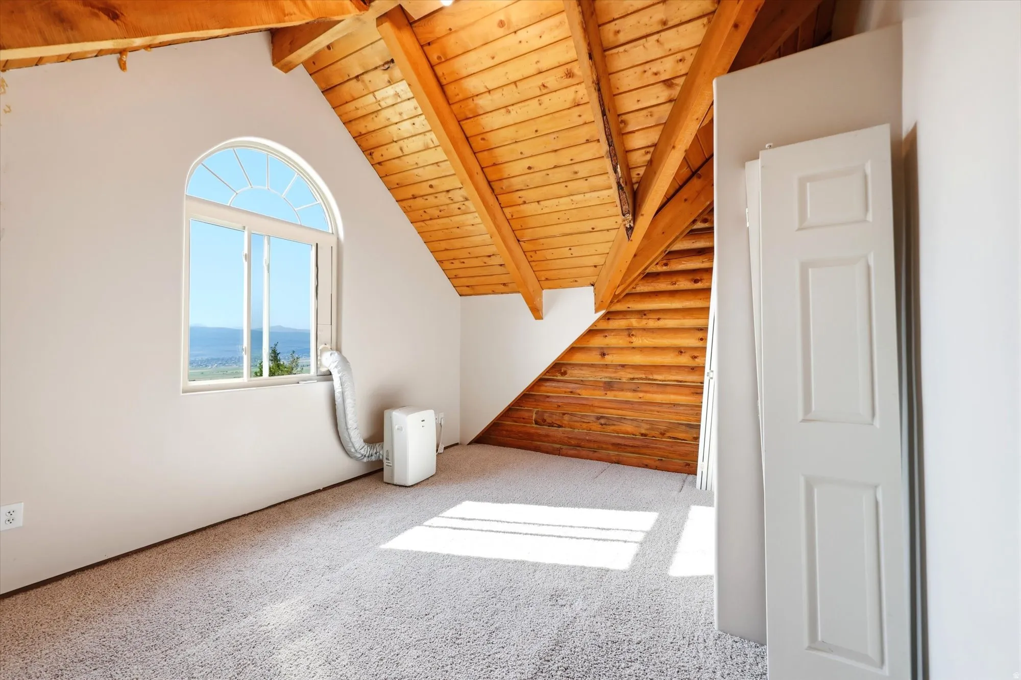 Carpeted attic space featuring exposed wood beams, a half-round arched window, and wood-paneled accent wall