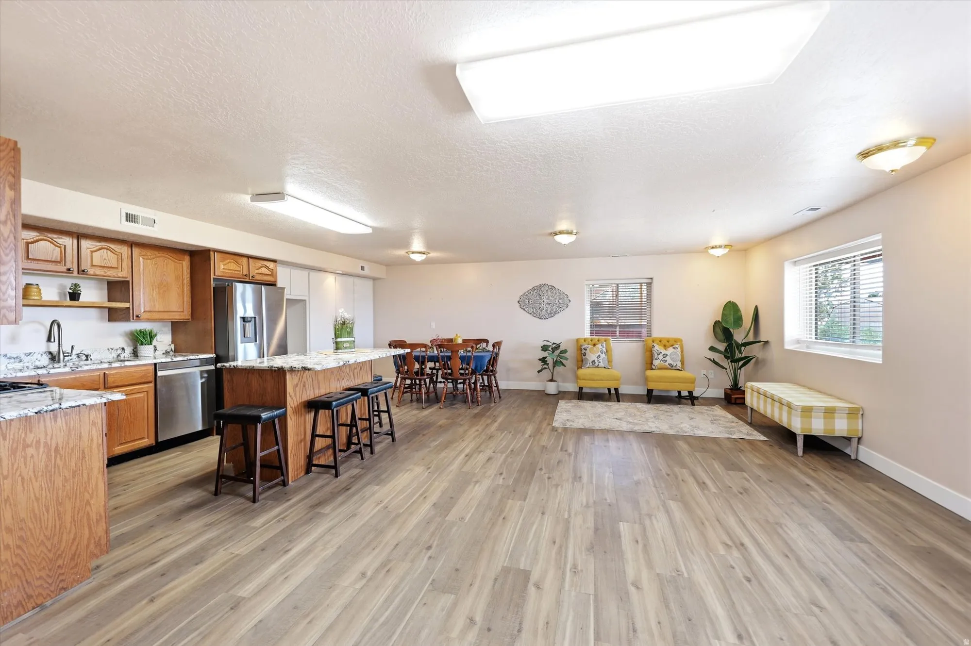 Open-concept living area featuring wood-finish flooring, a kitchen island with seating, stainless steel appliances, abundant overhead lighting, and multiple windows
