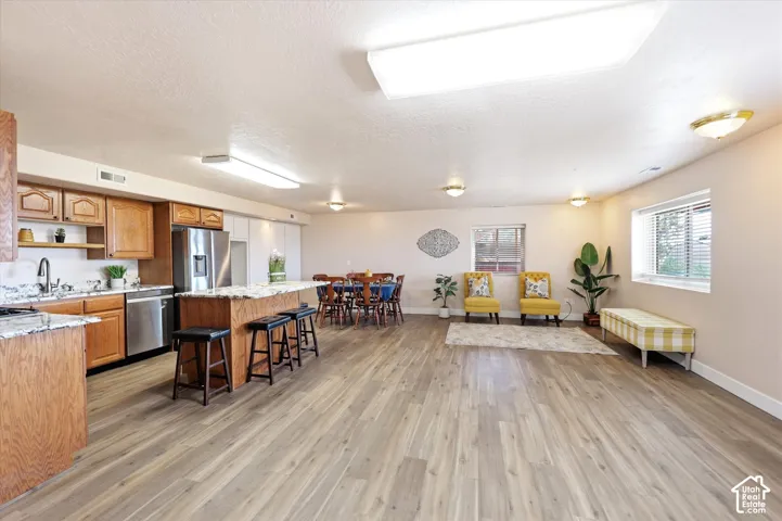 Open-concept living area featuring wood-finish flooring, a kitchen island with seating, stainless steel appliances, abundant overhead lighting, and multiple windows