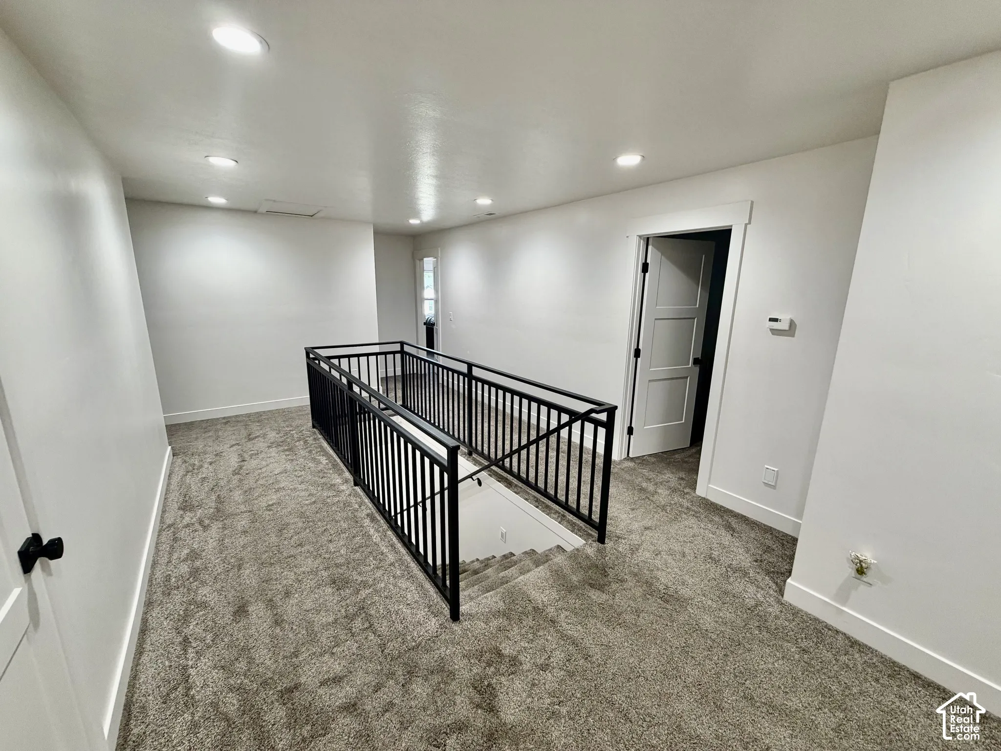 Upper-level landing featuring a modern black wrought iron railing system, recessed ceiling lighting, textured gray carpeting, a white three-panel interior door, and clean white baseboards