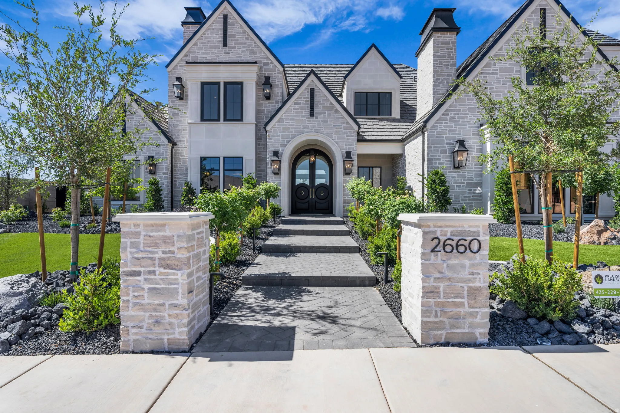Stone-clad exterior featuring multiple gables, dark-framed windows, a prominent arched entry, brick chimneys, and an inviting walkway