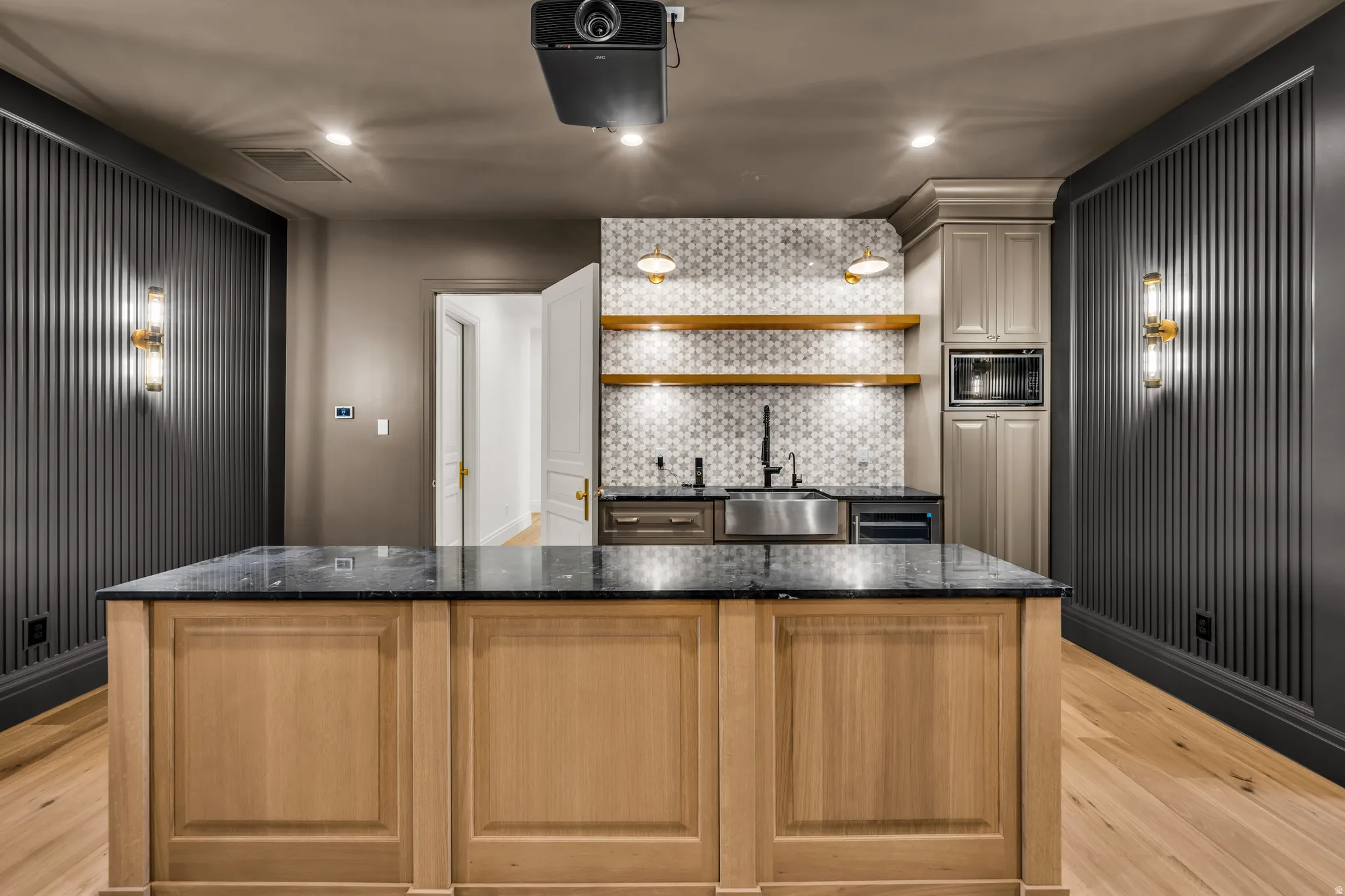 Custom entertainment space featuring a light wood-finish island with a dark stone countertop, a wet bar with patterned tile backsplash and open shelving, an integrated microwave, and wood-finish flooring