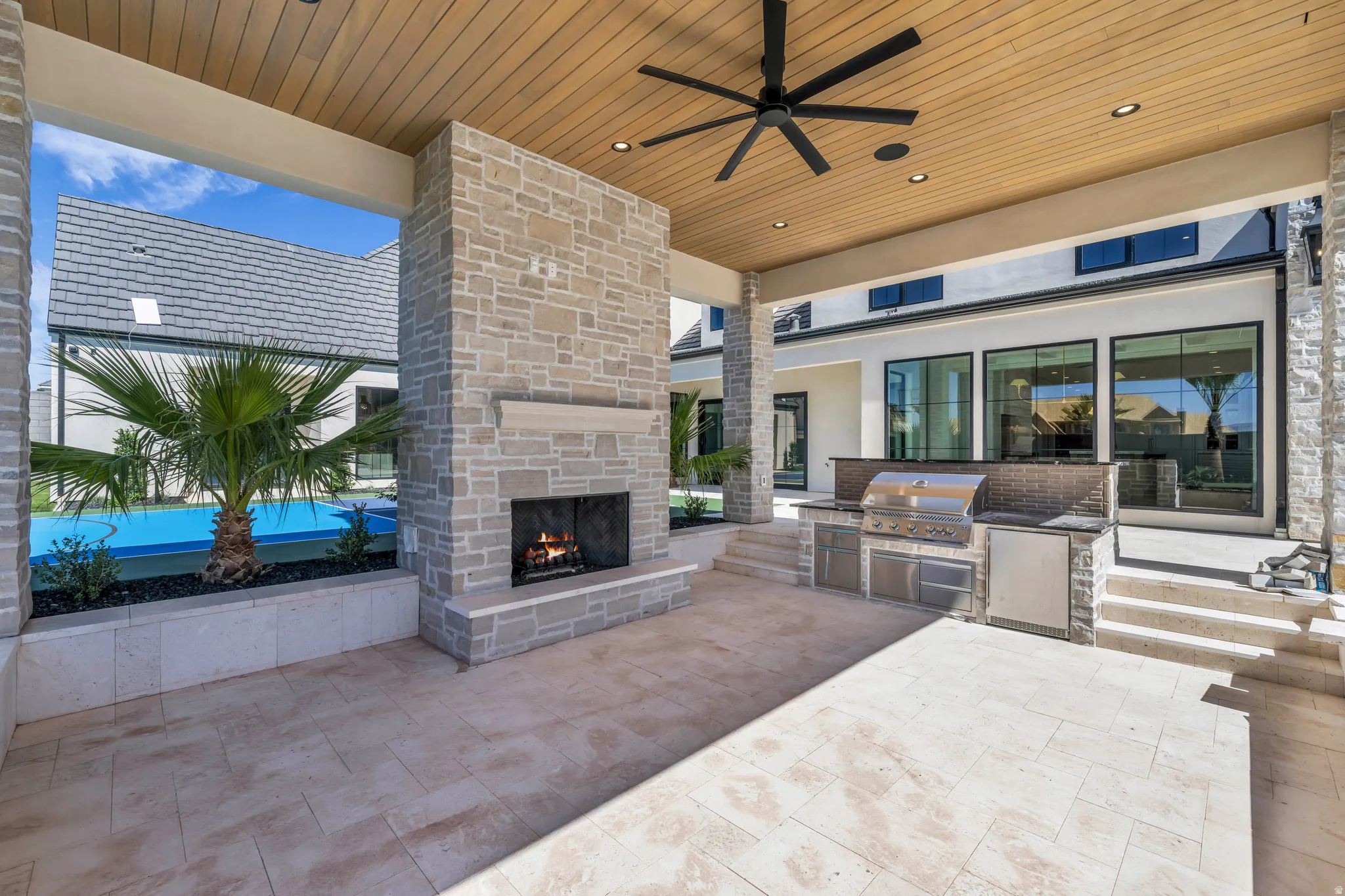 Expansive covered patio featuring a stone-clad fireplace with mantel, built-in outdoor kitchen with stainless steel appliances, wood-finish ceiling with recessed lighting and ceiling fan, and large format tile flooring