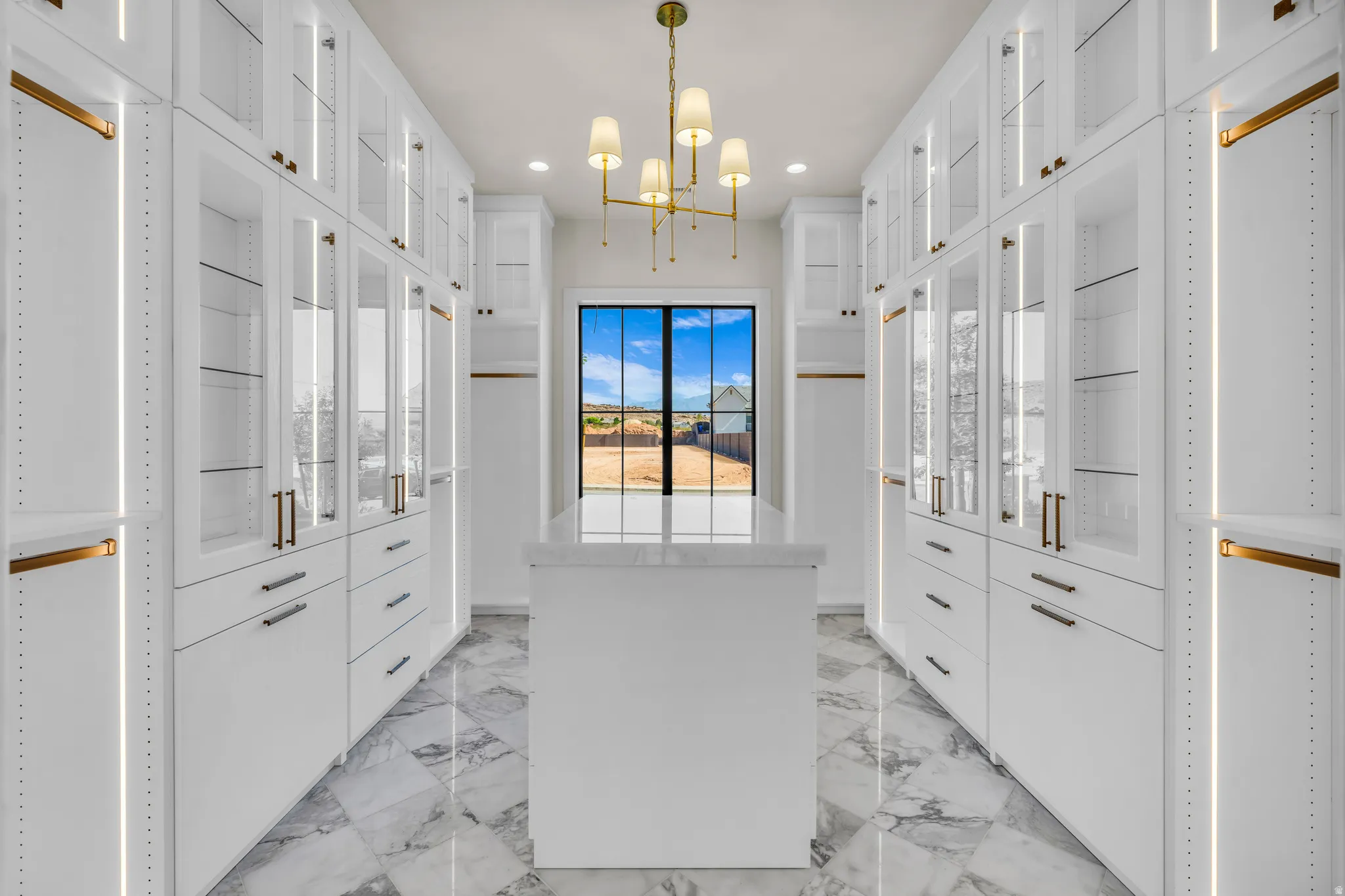 Spacious custom closet featuring extensive white cabinetry with glass-front uppers, an integrated center island, brass hardware, and interior lighting