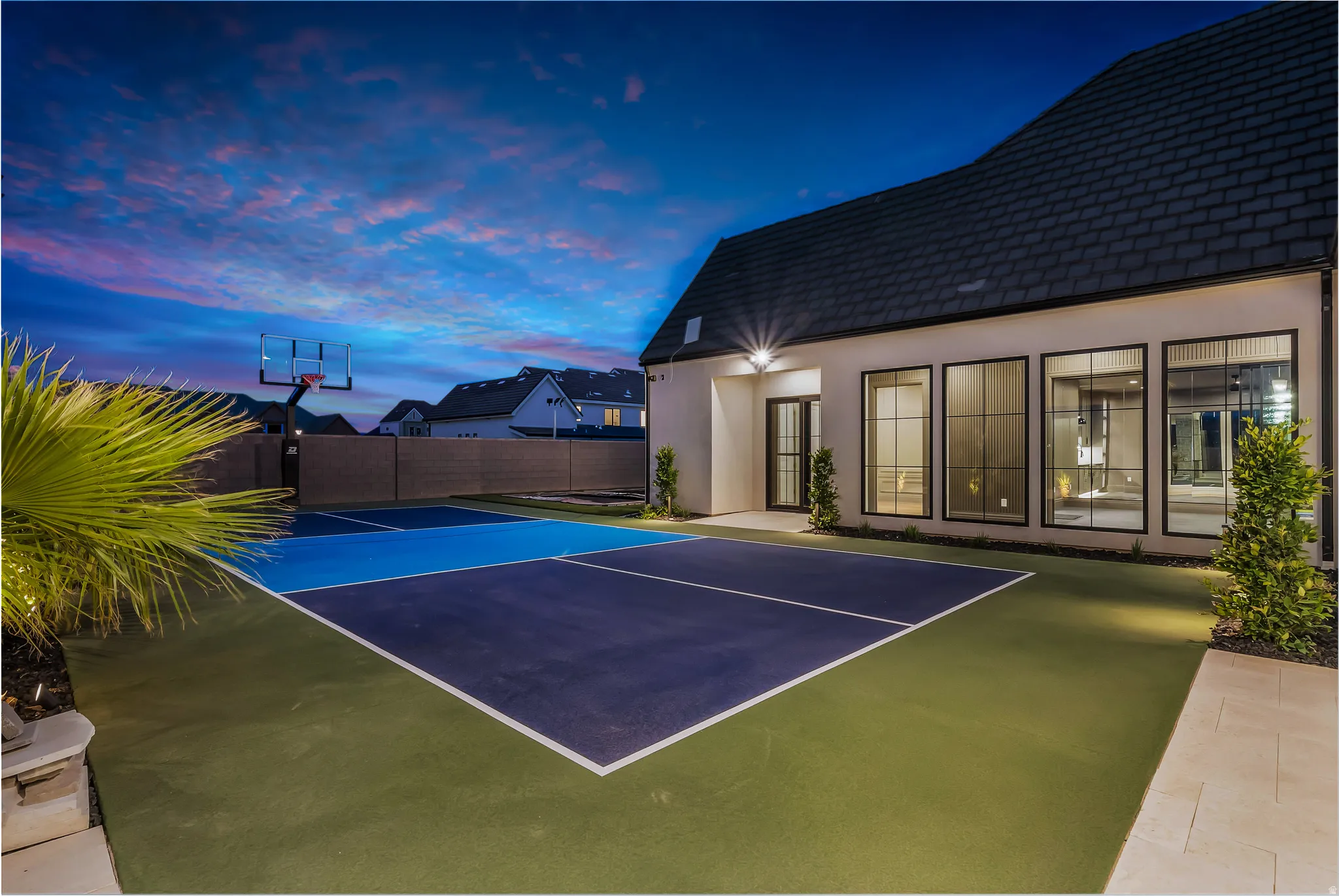 Multi-sport court with basketball hoop, surrounded by a modern stucco exterior with multiple large black-framed windows and a dark shingle roof