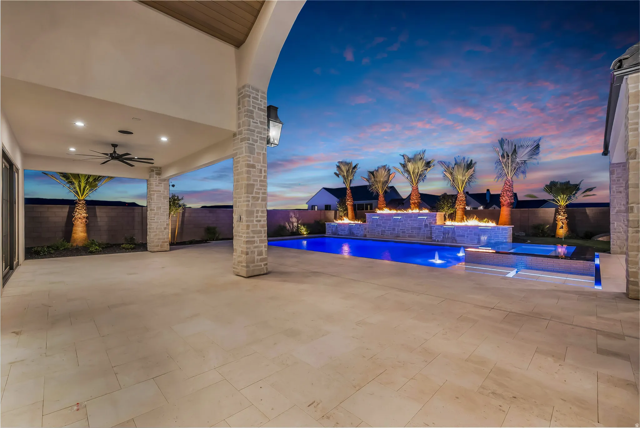 Expansive outdoor living area featuring a covered patio, a swimming pool with integrated spa, a decorative water feature with fire elements, and mature palm trees