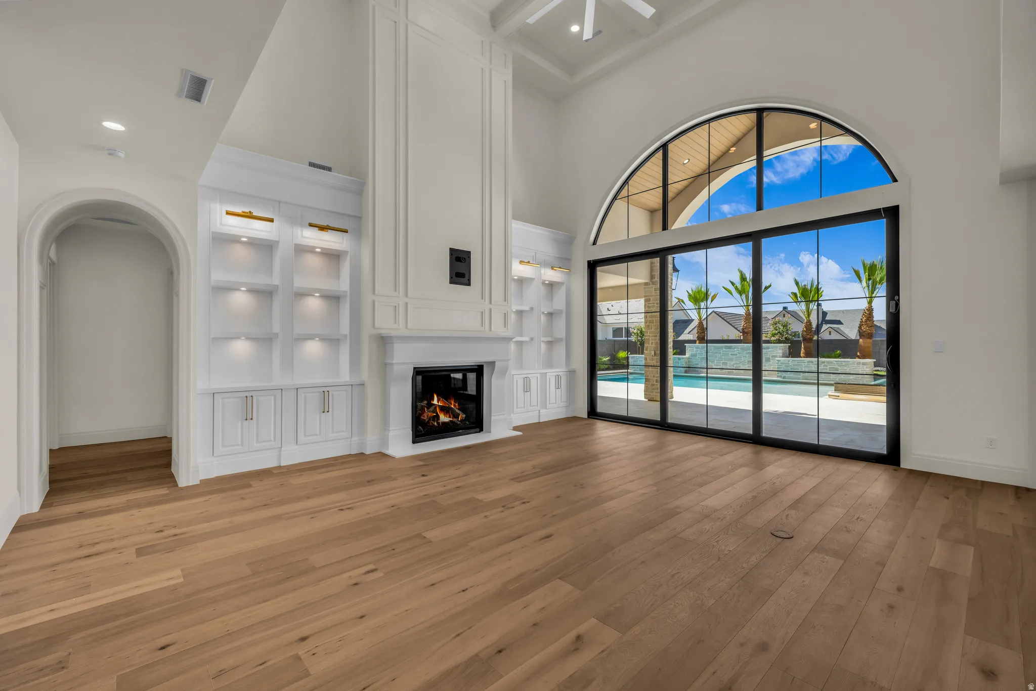 Great room featuring high ceilings, wood-finish flooring, a built-in fireplace with a mantel, integrated shelving with accent lighting, and an arched window wall with sliding glass doors