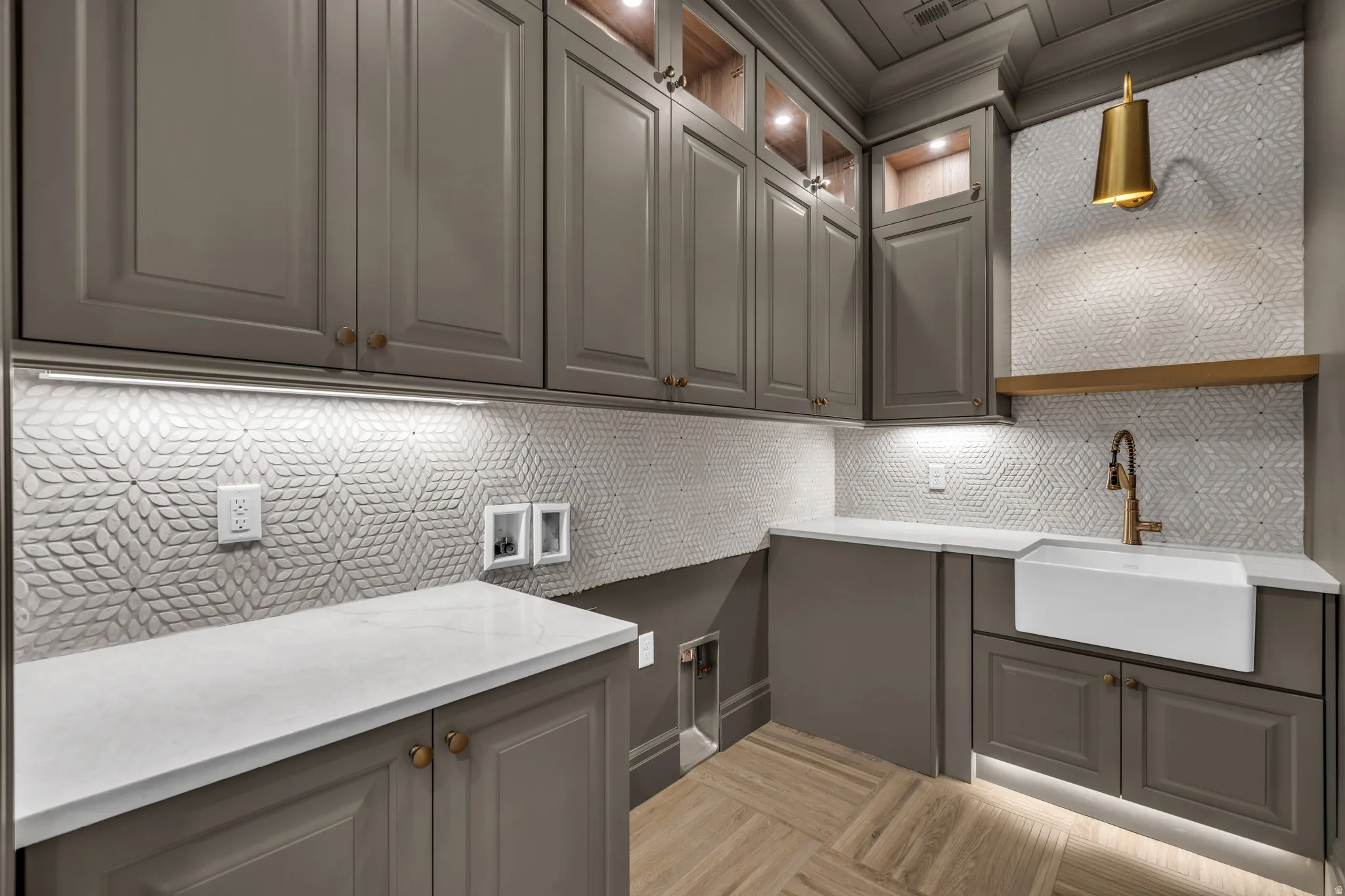 Laundry room featuring light gray cabinetry with gold-toned hardware, white quartz countertops, and a white farmhouse sink with a brass faucet