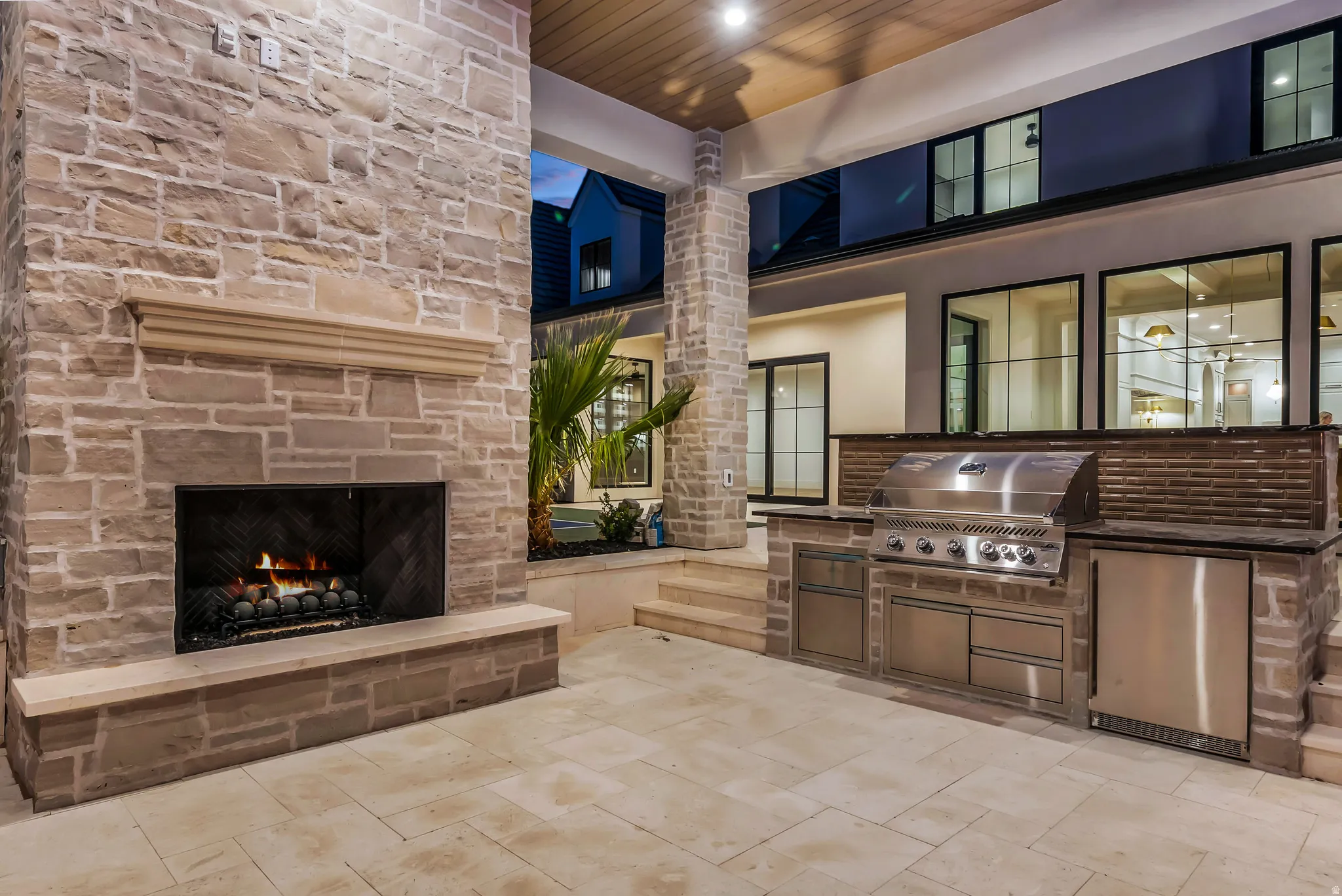 Expansive covered patio featuring a stone-clad outdoor fireplace with hearth, a built-in outdoor kitchen with stainless steel grill and refrigerator, and large format stone tile flooring