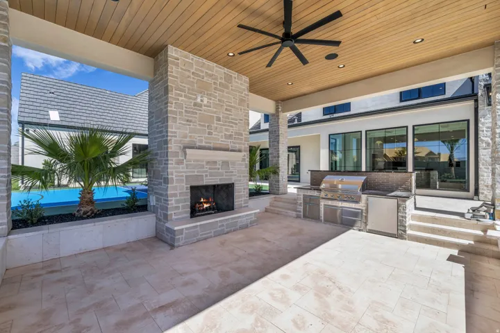Expansive covered patio featuring a stone-clad fireplace with mantel, built-in outdoor kitchen with stainless steel appliances, wood-finish ceiling with recessed lighting and ceiling fan, and large format tile flooring