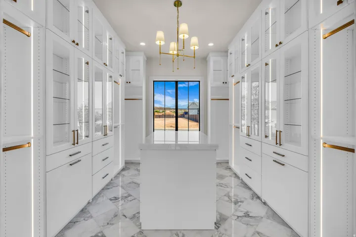 Spacious custom closet featuring extensive white cabinetry with glass-front uppers, an integrated center island, brass hardware, and interior lighting