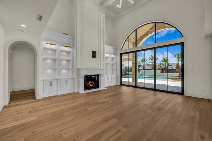Great room featuring high ceilings, wood-finish flooring, a built-in fireplace with a mantel, integrated shelving with accent lighting, and an arched window wall with sliding glass doors
