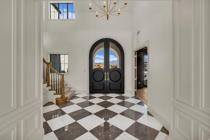 Grand entry foyer featuring checkerboard tile flooring, a prominent arched double-door entry, and a high ceiling with a gold-tone chandelier