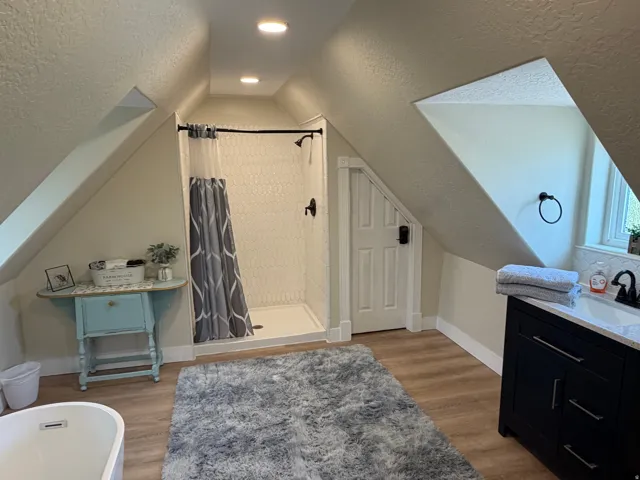 Bathroom featuring a walk-in shower with white hexagonal tile, matte black shower fixtures, wood-finish flooring, a freestanding bathtub, and a dark wood vanity with a light-colored countertop