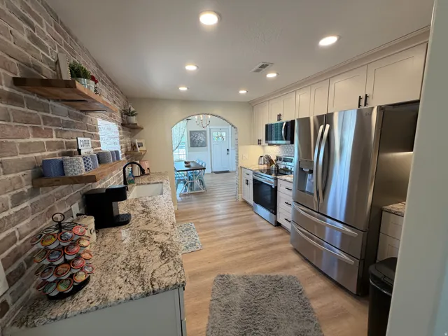 Modern kitchen featuring granite countertops, wood-finish flooring, white shaker cabinetry, stainless steel appliances, and a brick accent wall with floating shelves