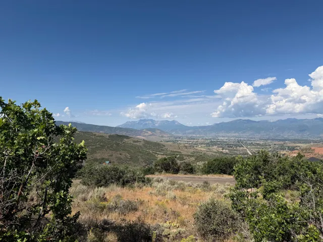 Expansive landscape featuring distant mountains, a broad valley, and a clear blue sky with scattered clouds