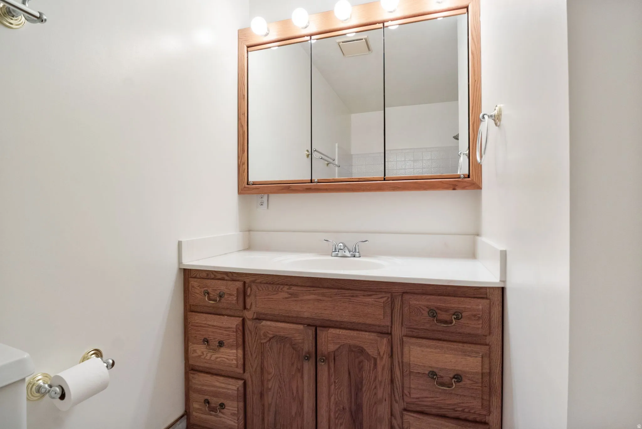 Wood-finish vanity with a white countertop and integrated sink, complemented by a tri-fold medicine cabinet with overhead vanity lighting