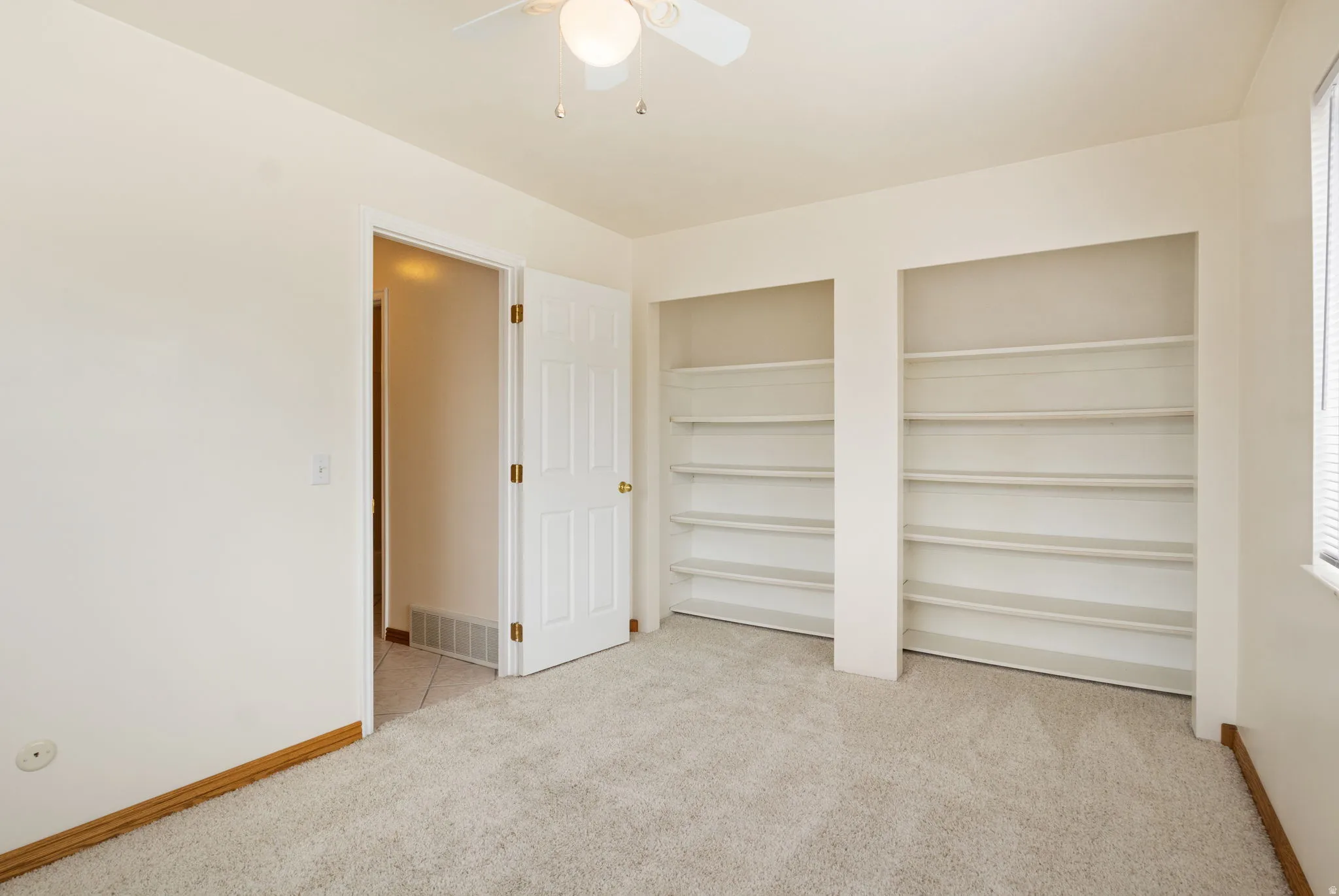 Carpeted room featuring two built-in shelving units, a ceiling fan with integrated lighting, and a window with blinds