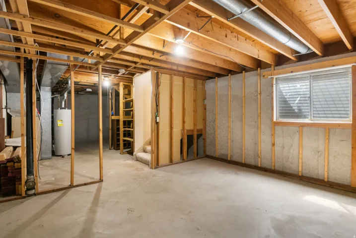 Unfinished basement featuring exposed wood framing, concrete flooring, a water heater, a window with horizontal blinds, and a partial staircase