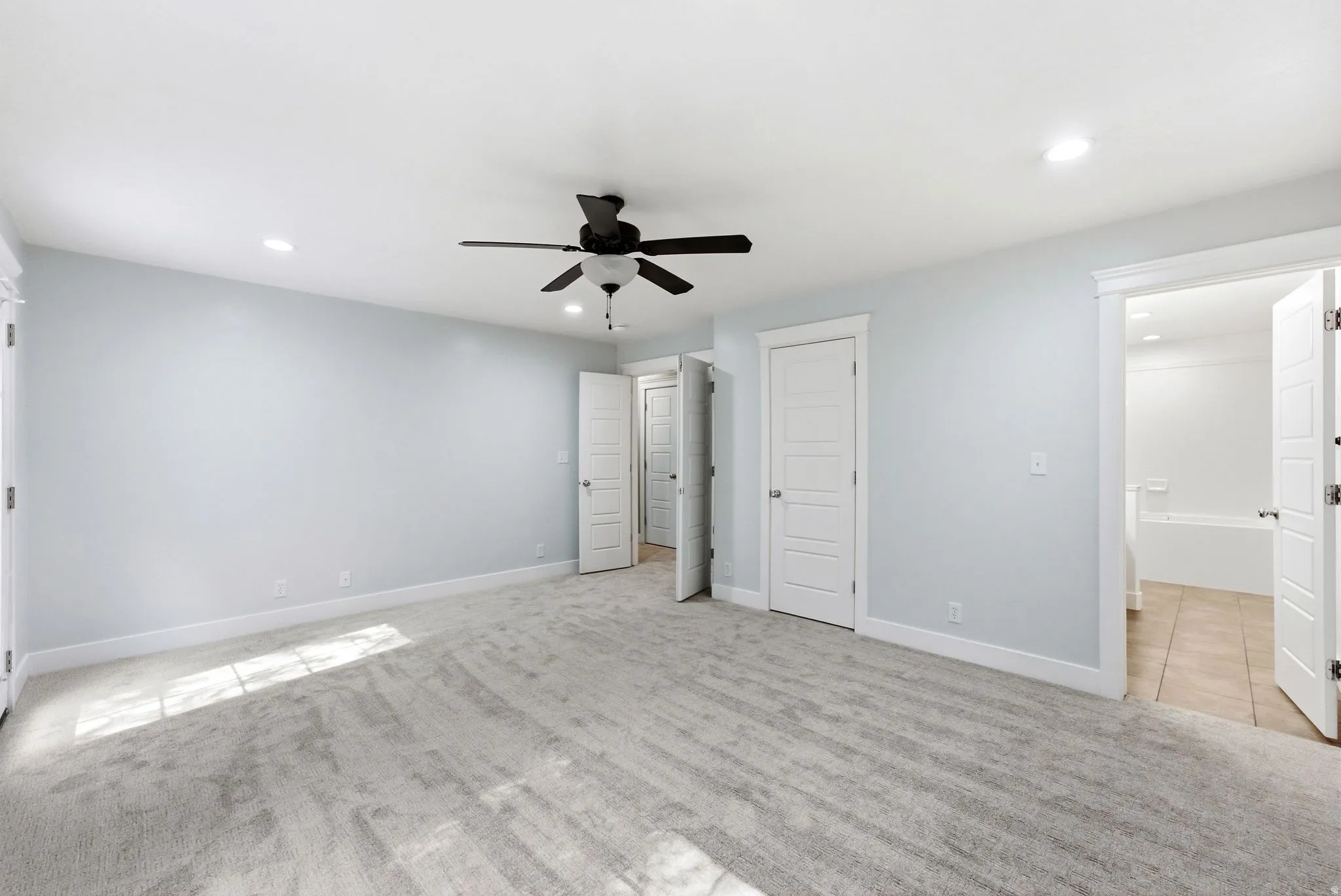 Spacious room featuring light gray carpeting, recessed lighting, a modern ceiling fan, and multiple white paneled doors