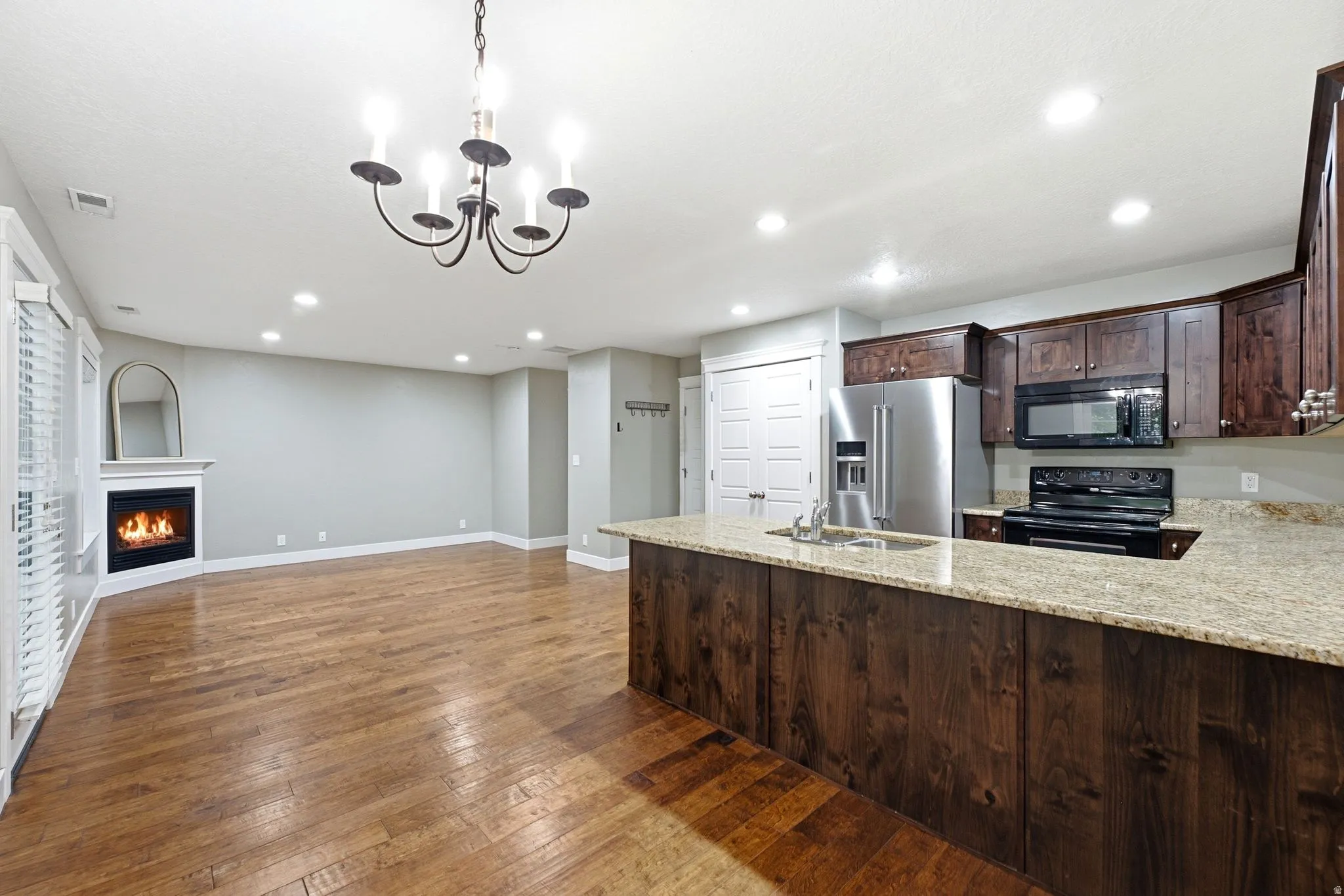 Open concept living space featuring wood-finish flooring, a corner-set fireplace with a white mantel, and a kitchen island with stone countertops