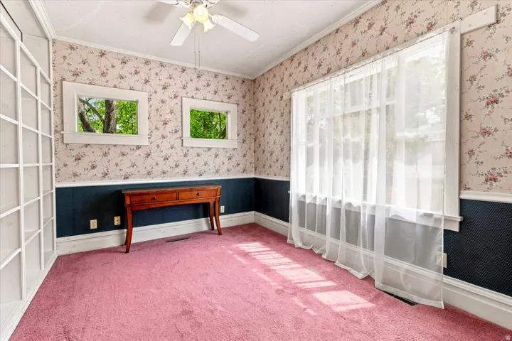 Room featuring floral wallpaper, white built-in shelving, two upper windows, a large window with sheer white curtains, and a ceiling fan
