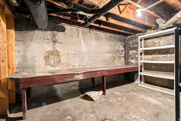 Unfinished basement featuring exposed concrete walls and flooring, visible ceiling joists, and overhead utility lines