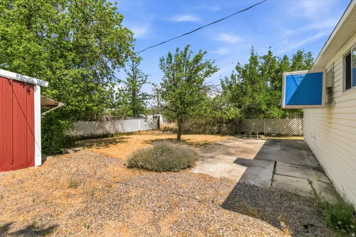 Expansive rear yard featuring a red storage shed, mature trees, a white perimeter fence, and concrete patio areas