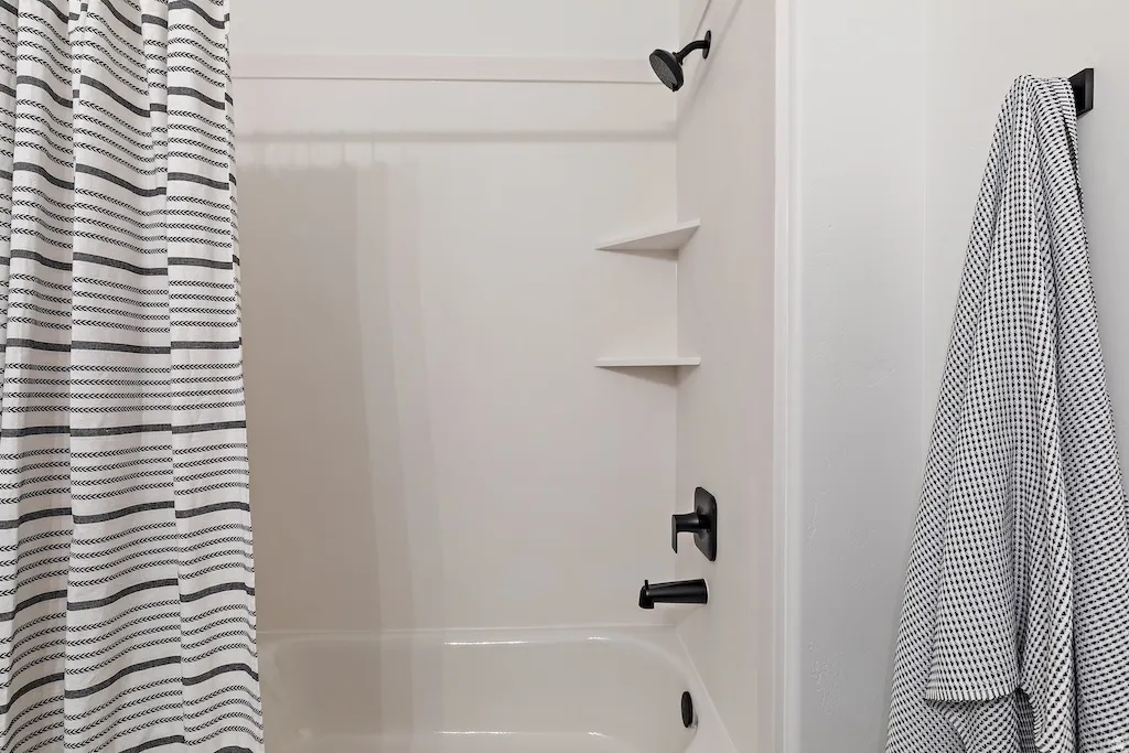 White tub/shower combination featuring built-in corner shelving, matte black fixtures, and a coordinating wall-mounted towel hook