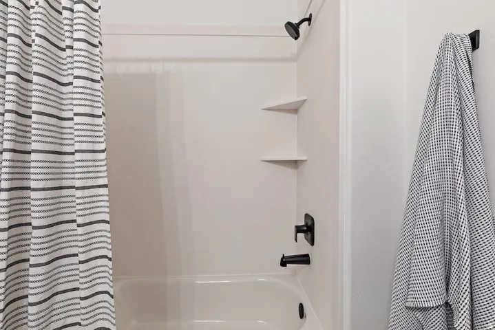 White tub/shower combination featuring built-in corner shelving, matte black fixtures, and a coordinating wall-mounted towel hook