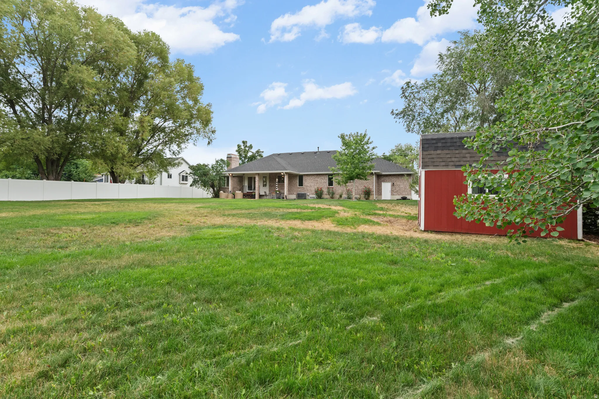 Spacious backyard featuring a manicured lawn, a detached red storage shed with a dark shingle roof, mature perimeter trees, and a white privacy fence