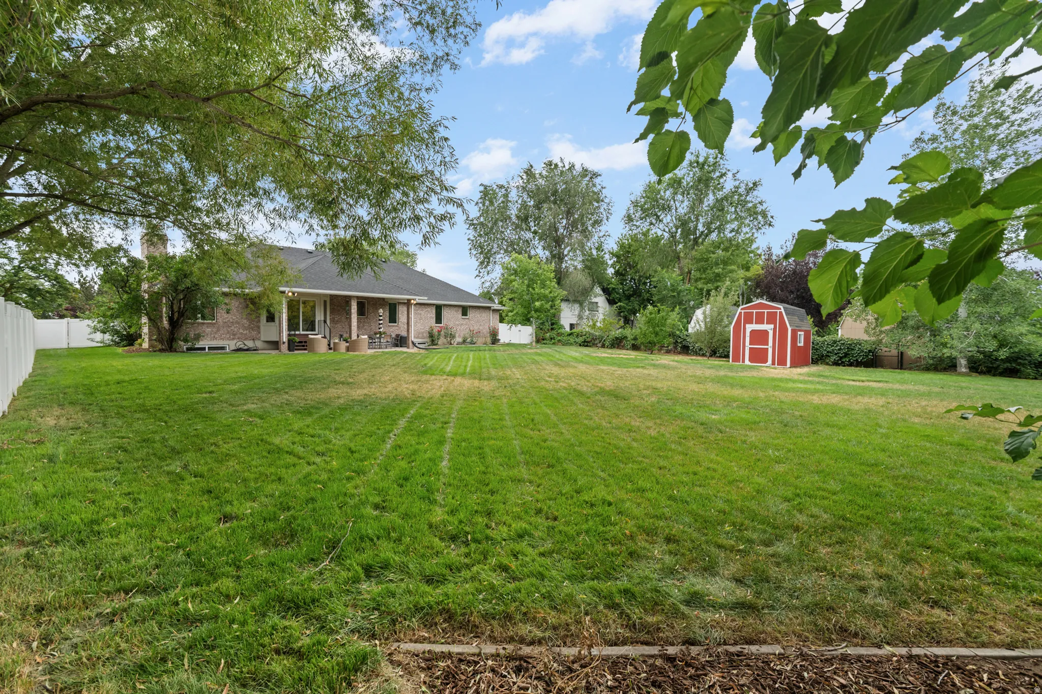 Expansive green lawn featuring a red storage shed, a white privacy fence, and mature trees