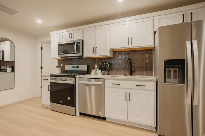 Modern kitchen featuring light wood-finish flooring, white cabinetry with matte black hardware, stainless steel appliances, textured backsplash, and solid surface countertops